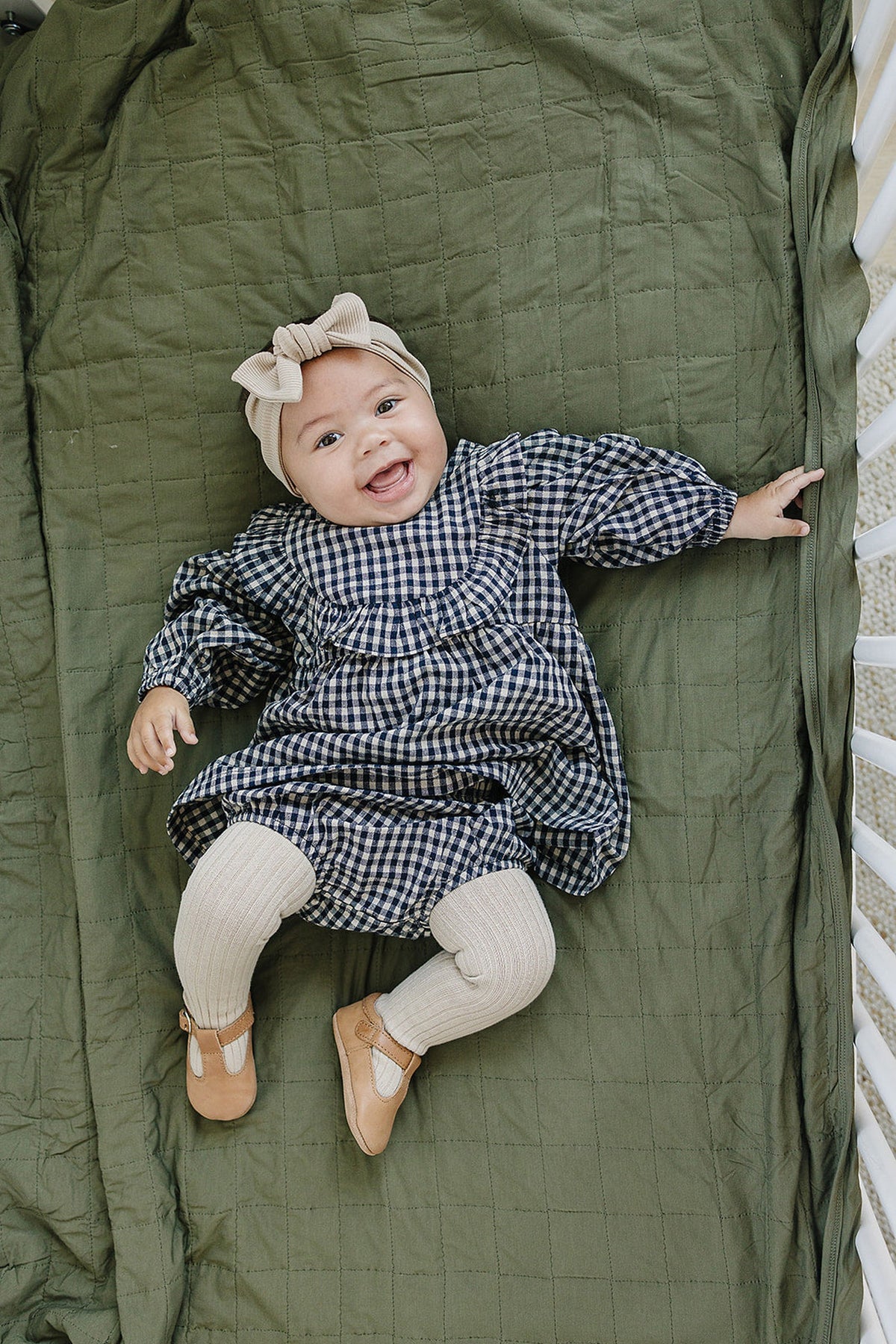 A baby wearing a blue checkered dress, beige ribbed tights, and brown shoes lies on a green quilt.