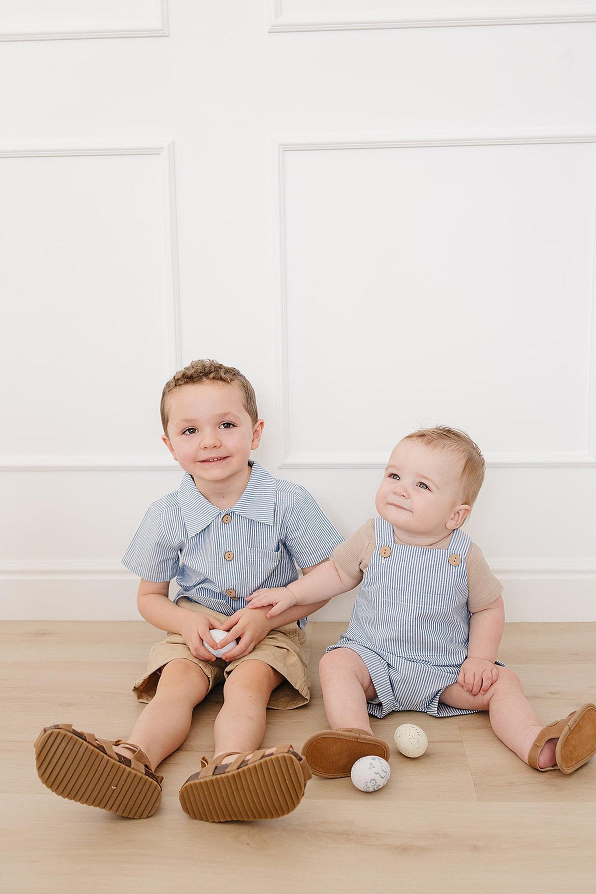 Two young children sit on a wooden floor, one wearing a blue striped button-up shirt and the other in a.