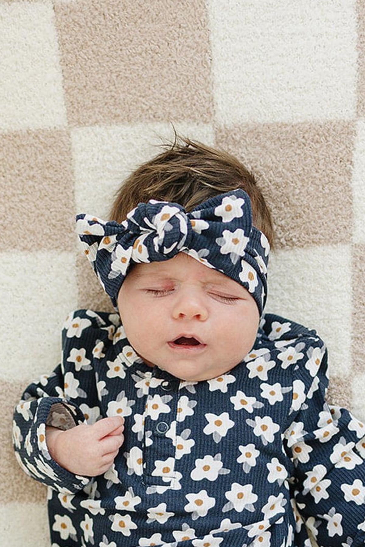 Baby wearing a navy daisy-patterned head wrap and matching outfit, lying on a checkered beige and white blanket.