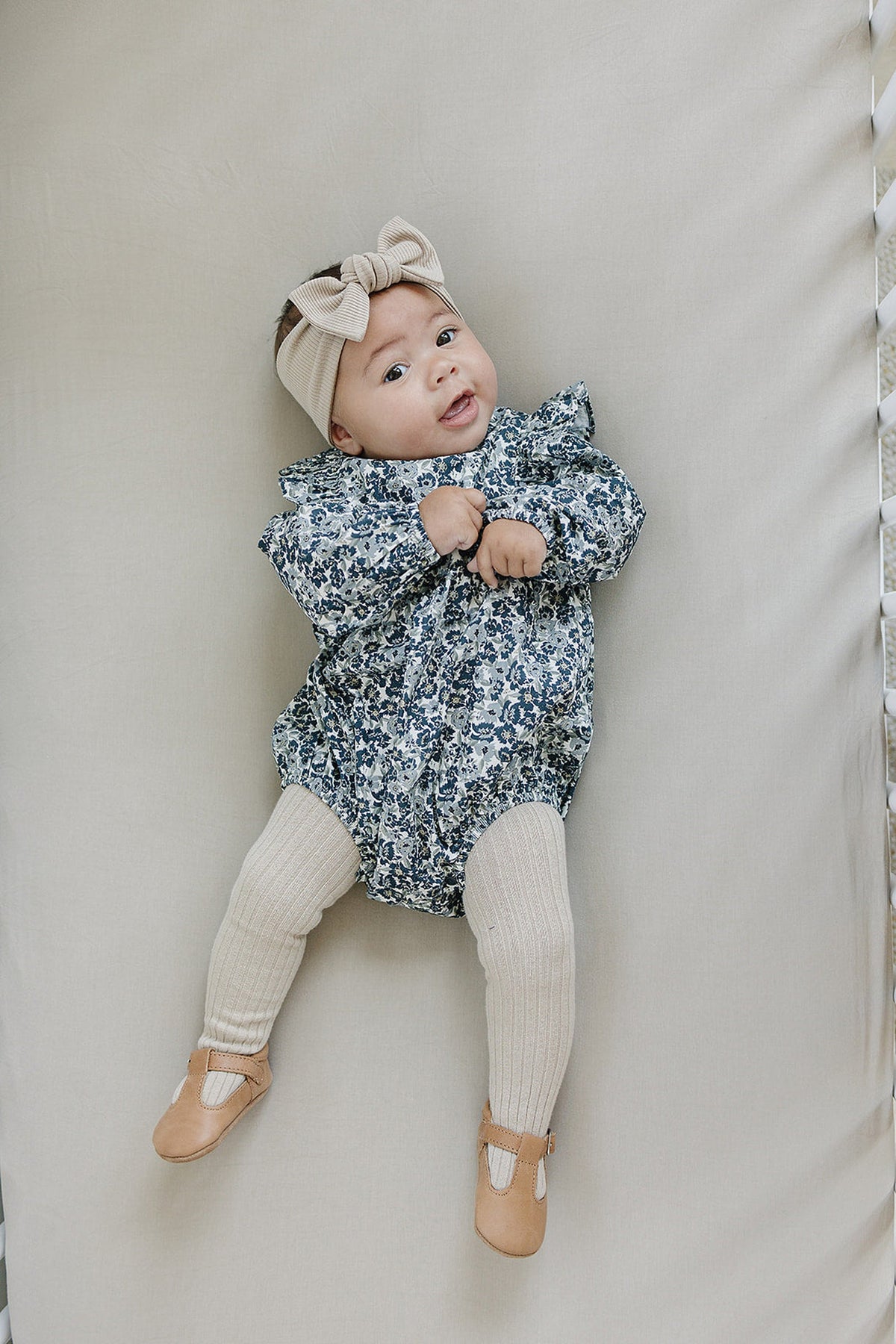 A baby wearing a floral romper, beige ribbed tights, tan shoes, and a matching headband lies on a cream surface.