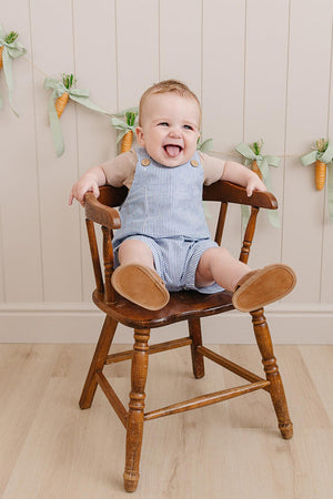 A smiling baby sits in a wooden chair wearing blue striped short overalls and tan shoes, with a decorative carrot.