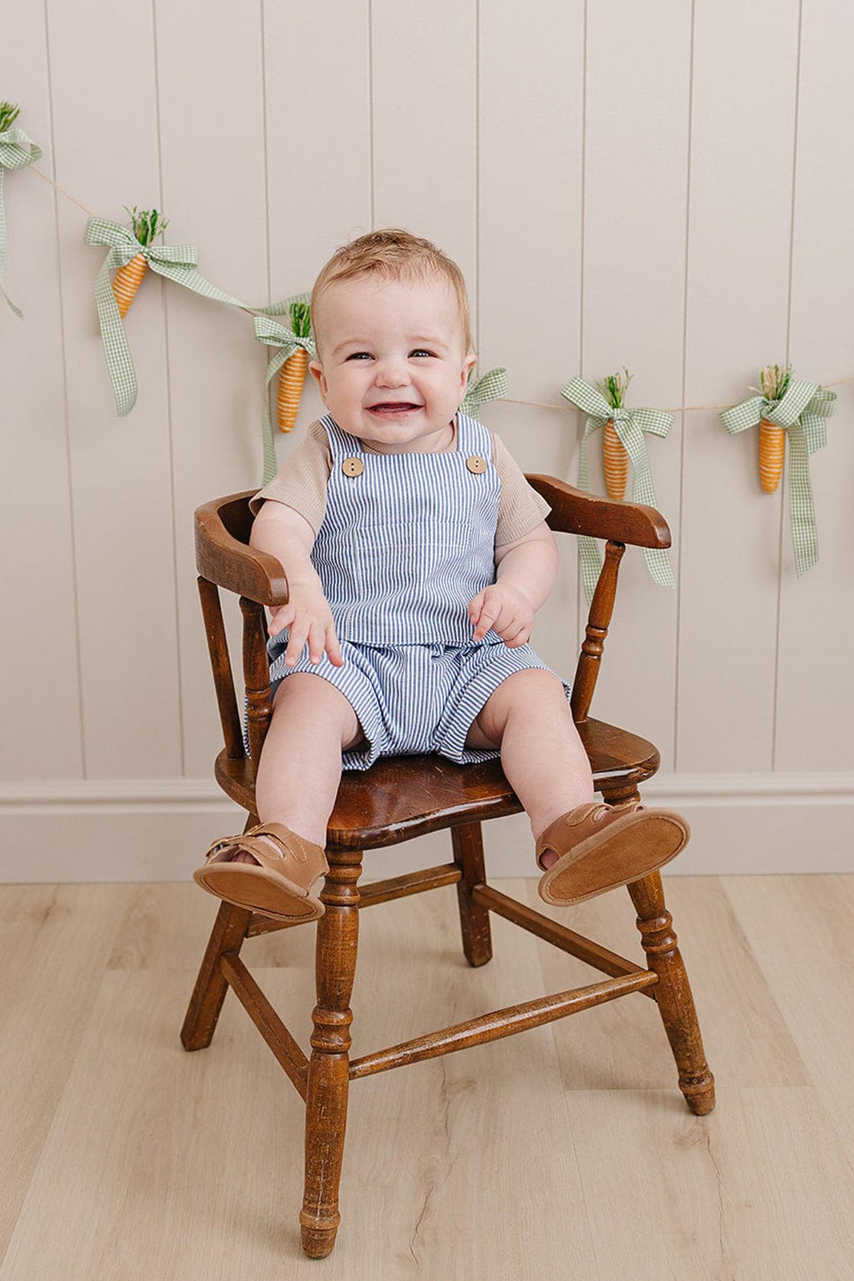 A smiling baby boy sits on a wooden chair, wearing blue striped short overalls and brown sandals, with a decorative.