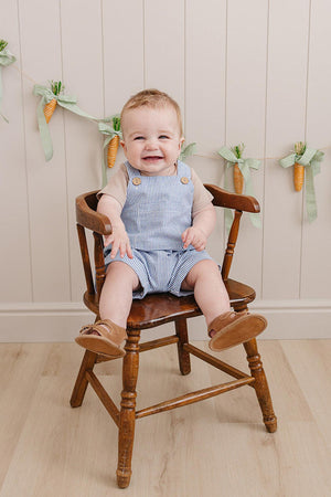 A smiling baby boy sits on a wooden chair, wearing blue striped short overalls and brown sandals, with a decorative.