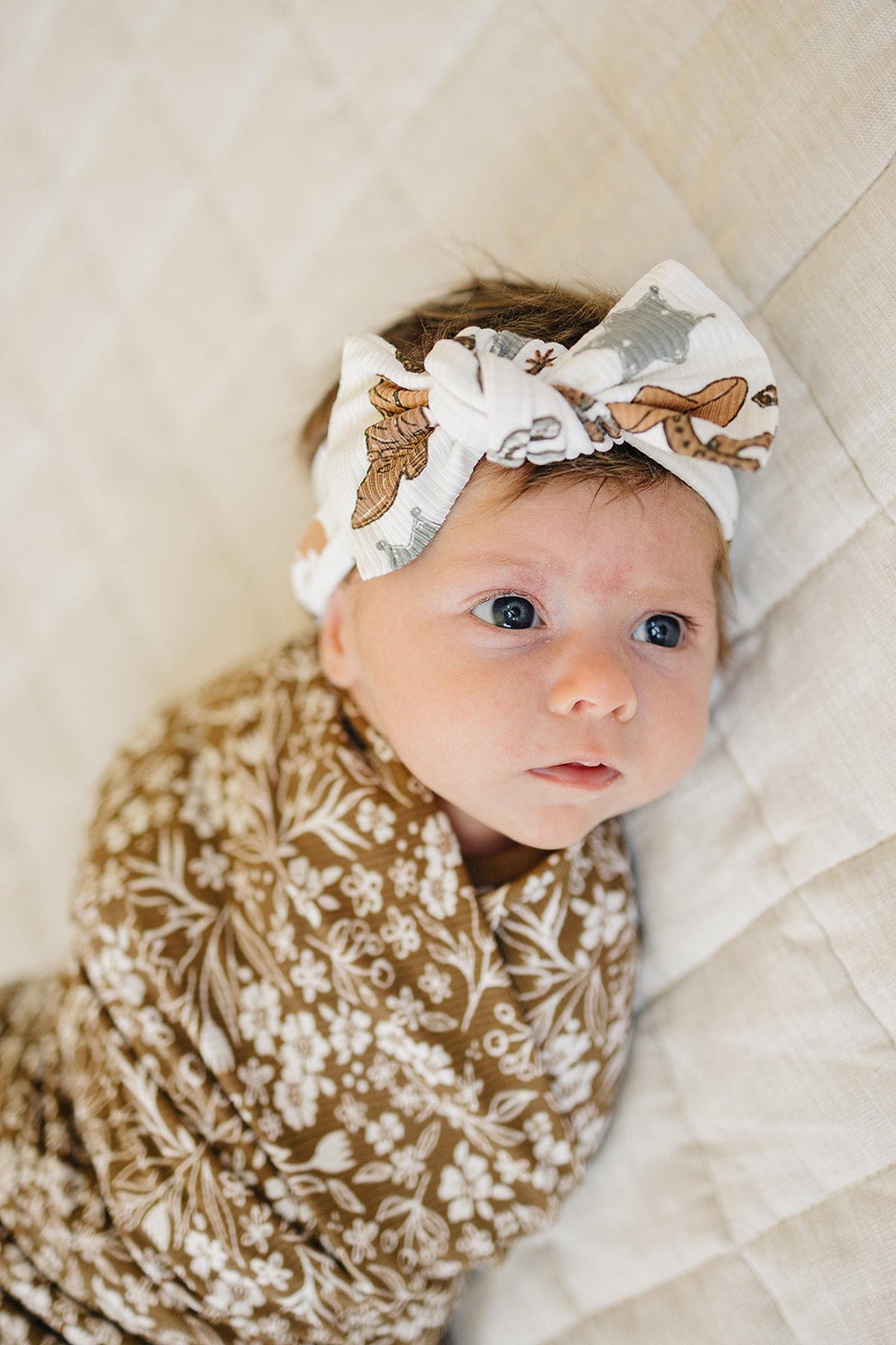 Baby wearing a white head wrap with cowboy print and a brown floral blanket, lying on a quilted surface.
