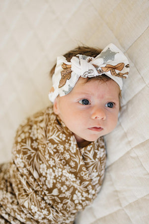 Baby wearing a white head wrap with cowboy print and a brown floral blanket, lying on a quilted surface.