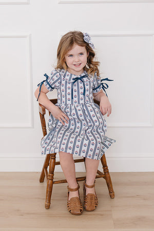 A young girl sits on a wooden chair, wearing a blue and white block print dress with a collar and ribbon ties. She smiles, accessorized with a matching hairpiece.