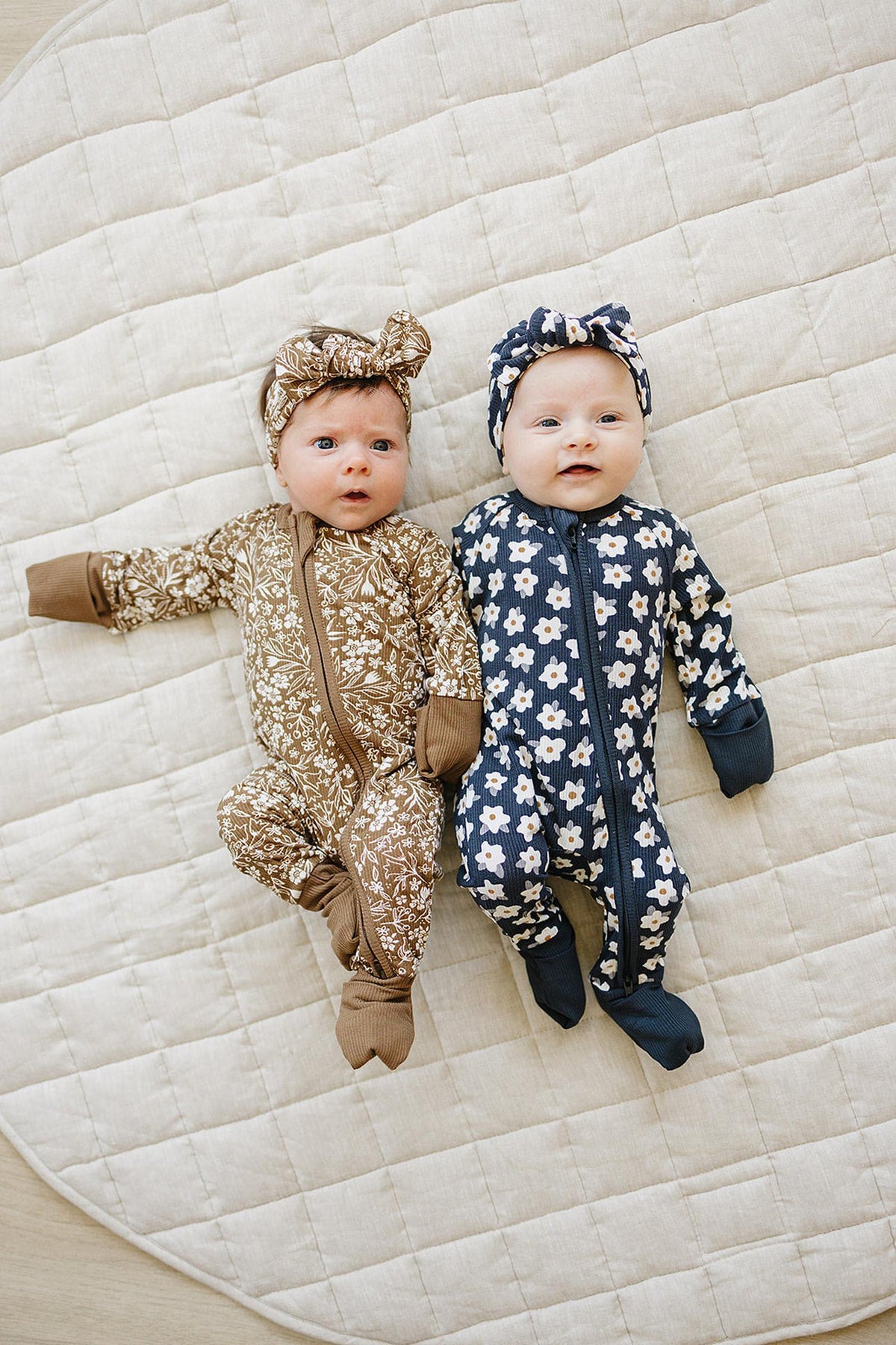Two babies lying on a quilt, wearing floral-patterned zipper pajamas and matching headbands, one brown and one navy.