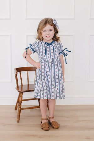 A young girl poses in a stylish blue and white block print dress with ribbon ties, complemented by a matching hair accessory.