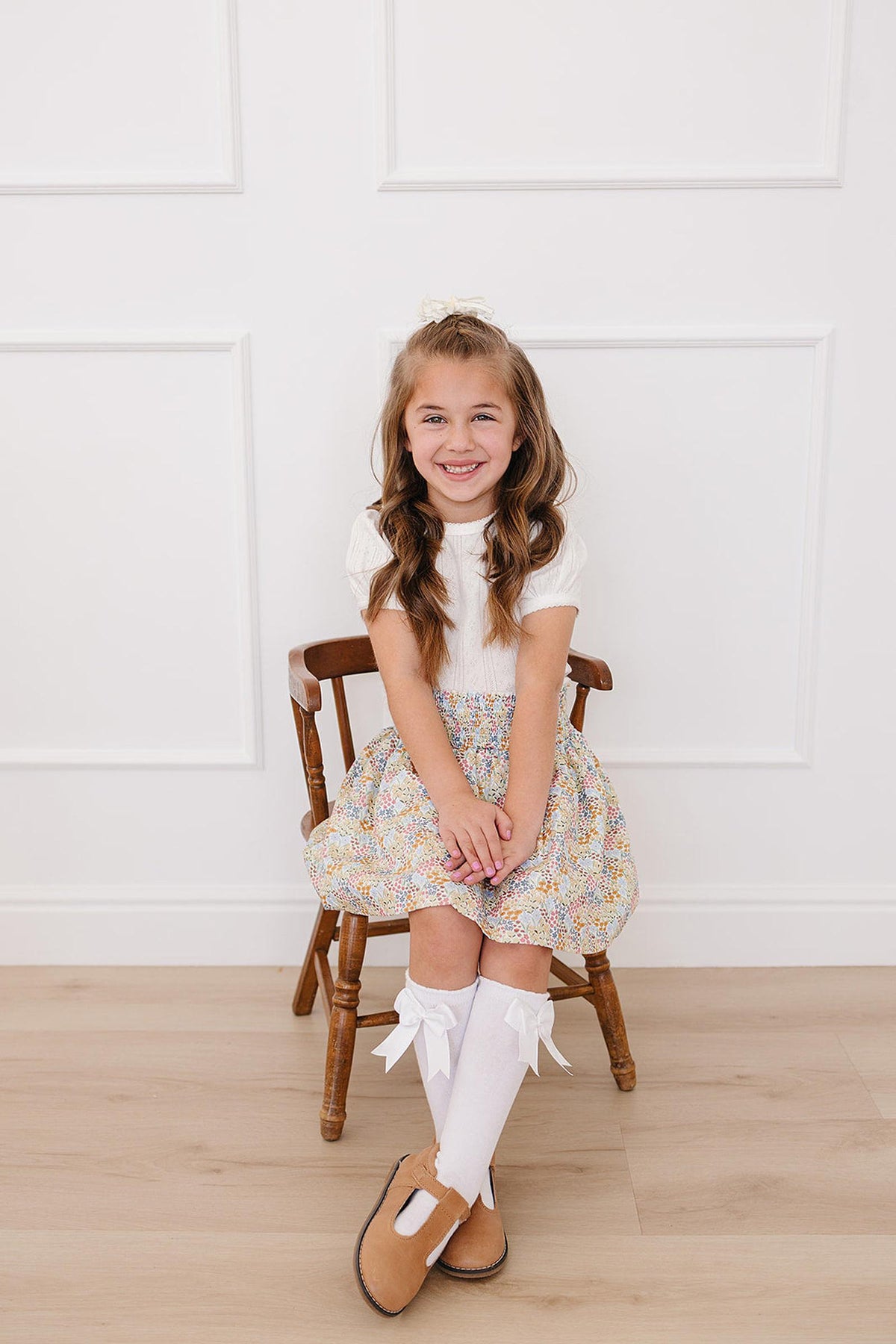 A smiling girl sits on a wooden chair, wearing a floral skirt, white top, and knee-high socks with bows.