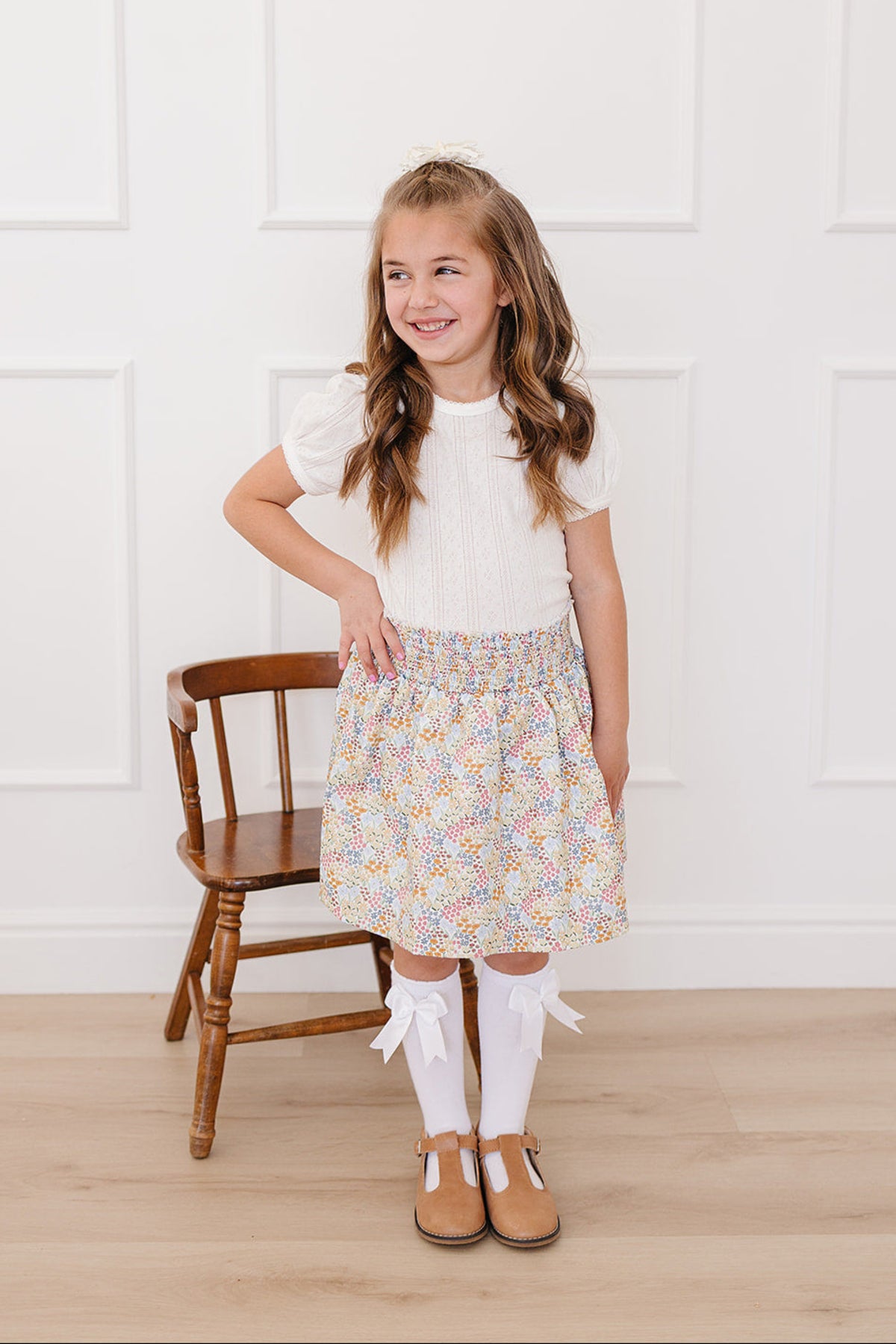 A smiling girl wears a floral cotton skirt with pastel colors, paired with a white top, knee-high socks, and brown.