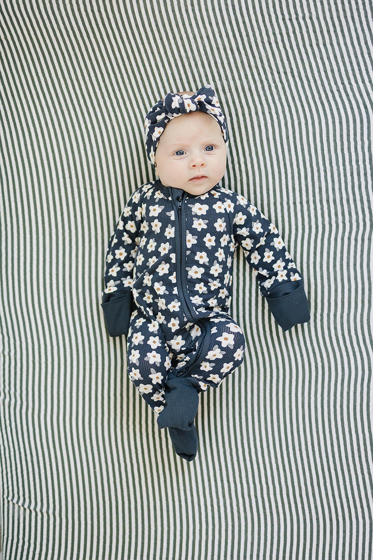 A baby in navy daisy-patterned pajamas and matching headband lies on a striped background.