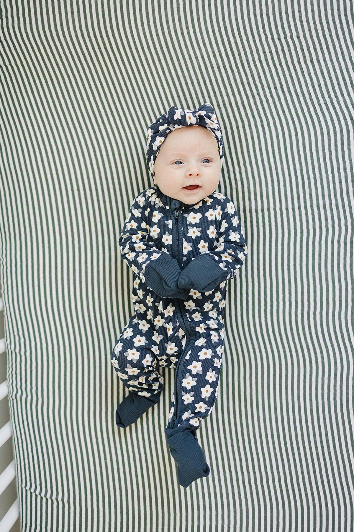 A baby in navy daisy-patterned pajamas with matching headband lies on a striped green and white blanket.