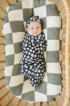 A baby swaddled in a navy daisy-patterned bamboo fabric lies on a green and white checkered blanket in a wicker.