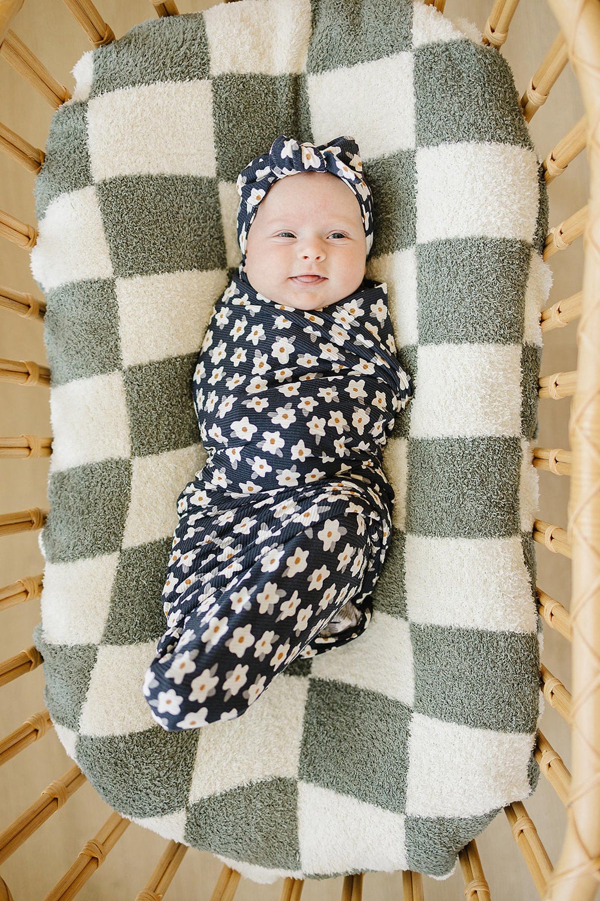 A smiling baby is swaddled in a navy daisy-patterned bamboo fabric on a checkered blanket in a basket.