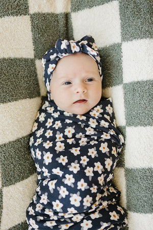 A baby wrapped in a navy daisy-patterned swaddle and matching headband lies on a textured checkered blanket.