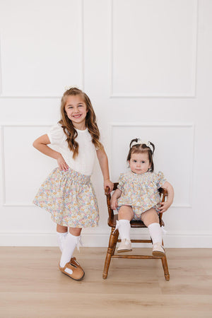 Two young girls model colorful floral outfits in a bright, airy room. The older girl wears a cheerful skirt, while.