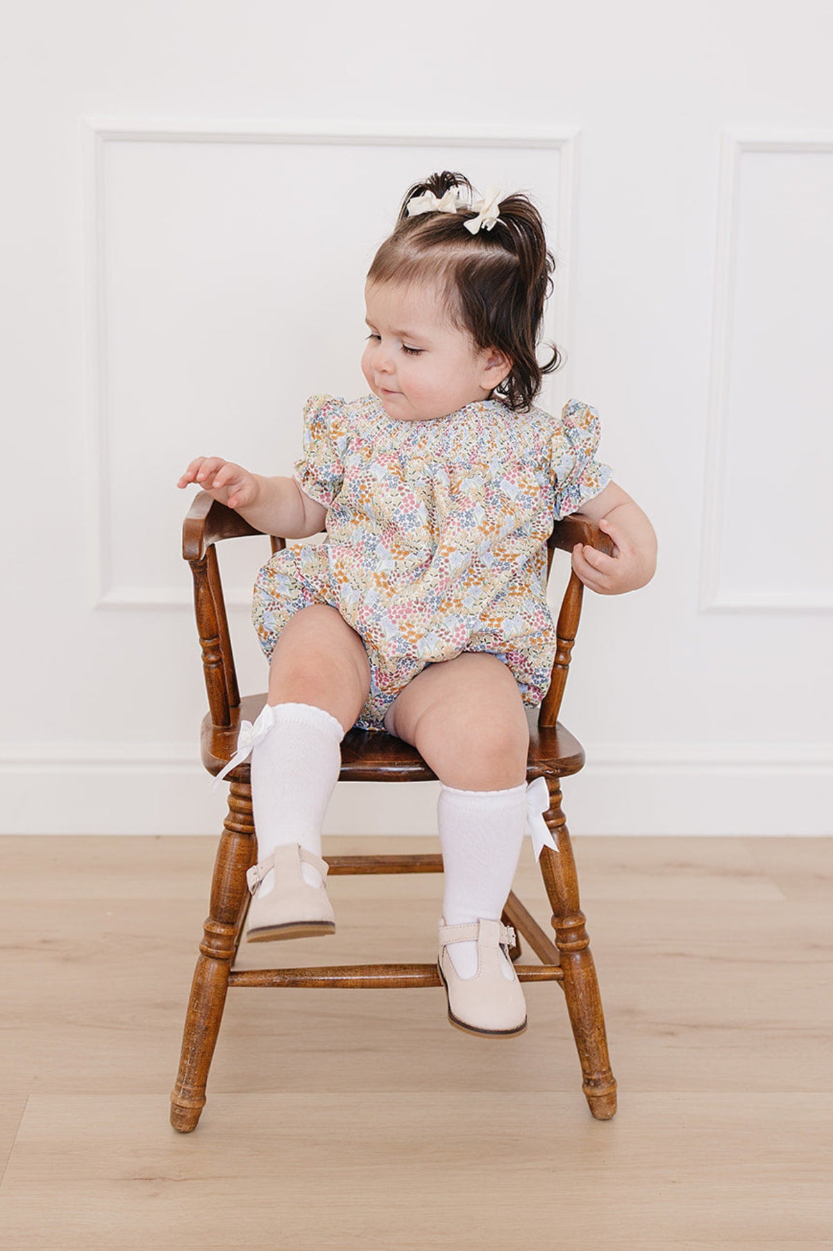 A baby girl sits on a wooden chair, wearing a floral romper with puff sleeves, white knee-high socks, and beige.
