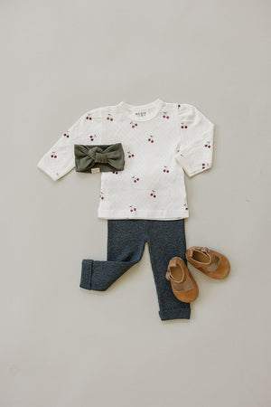 A white long sleeve T-shirt with cherry prints, paired with dark blue pants, a green headband, and brown shoes.