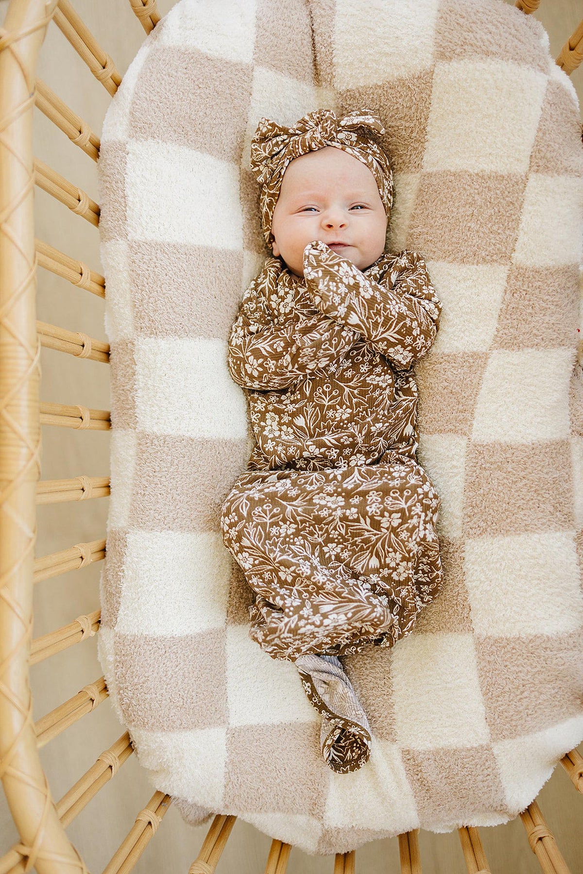 A baby wrapped in a floral brown swaddle and matching head wrap rests in a cozy, checkered bassinet. The soft fabrics provide a gentle touch for delicate skin.