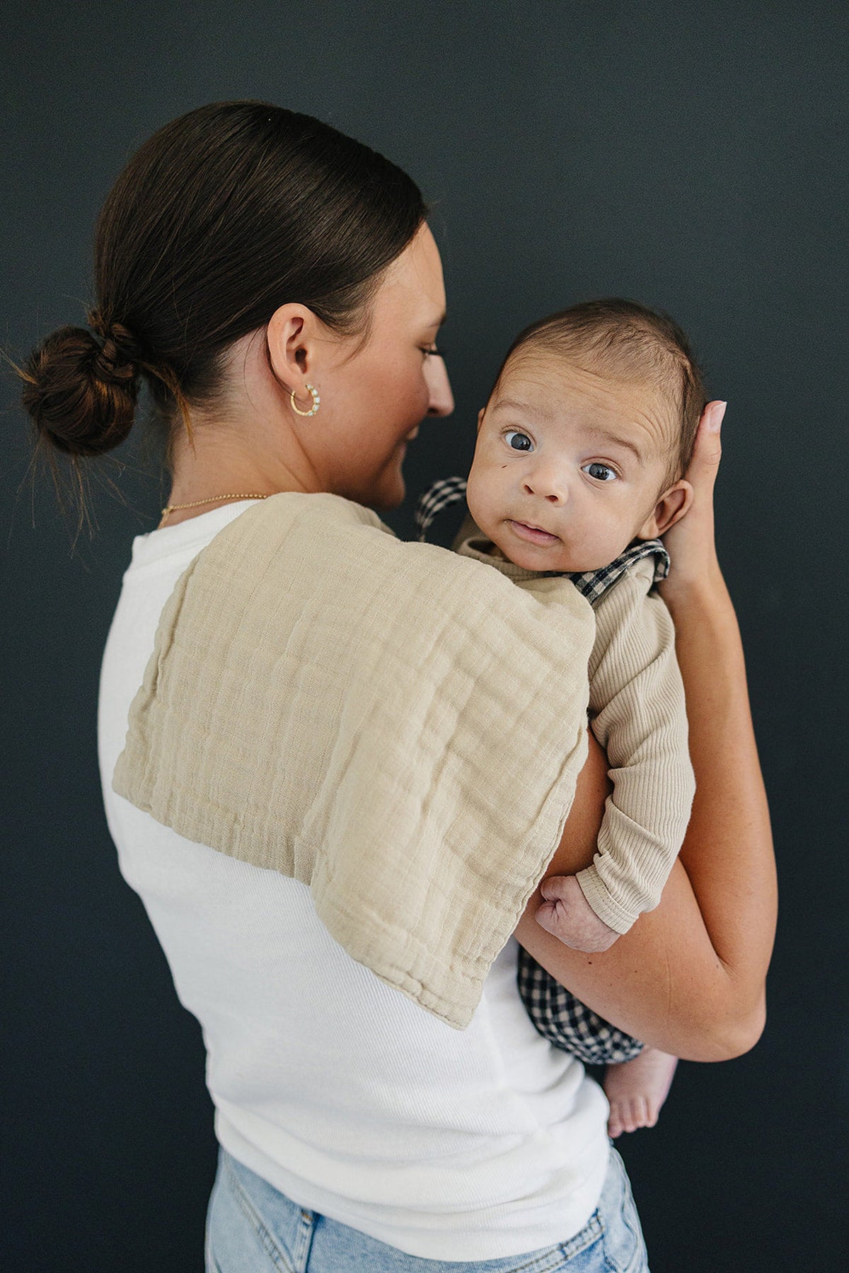 A woman holds a baby against her shoulder, draped with a soft, oatmeal-colored muslin burp cloth. The background is dark.