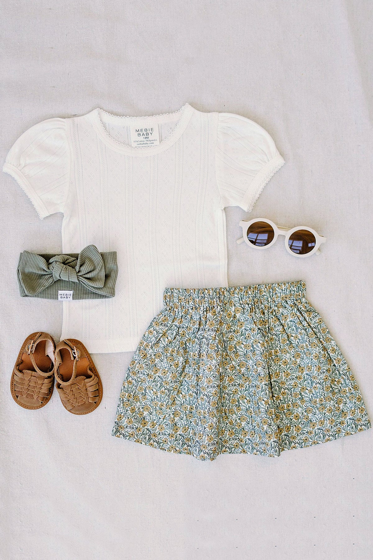 White pointelle t-shirt with puff sleeves, floral skirt, brown sandals, green bow headband, and white sunglasses on fabric.