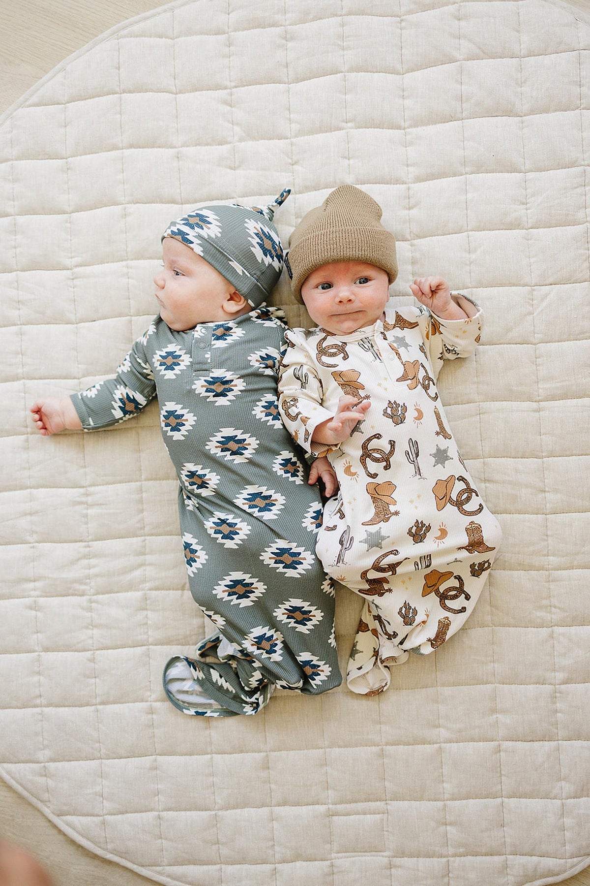 Two babies lie on a quilted mat, wearing patterned gowns; one in green with geometric designs, the other in cowboy-themed.