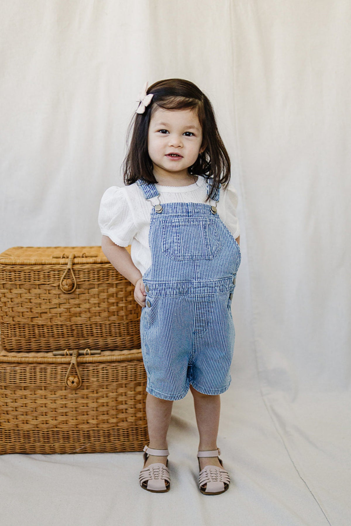 A child wearing blue striped overalls and a white shirt stands next to wicker baskets on a neutral background.