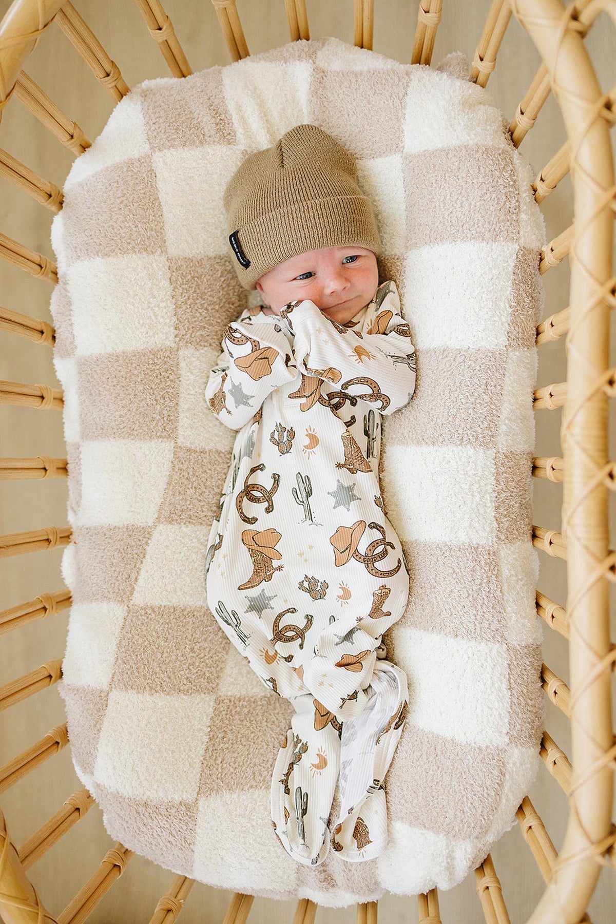 A baby in a cowboy-themed gown and beige hat lies in a checkered, cushioned wicker crib.