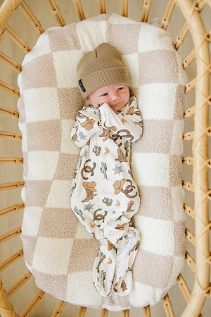 A baby in a cowboy-themed gown and beige hat lies in a checkered, cushioned wicker crib.