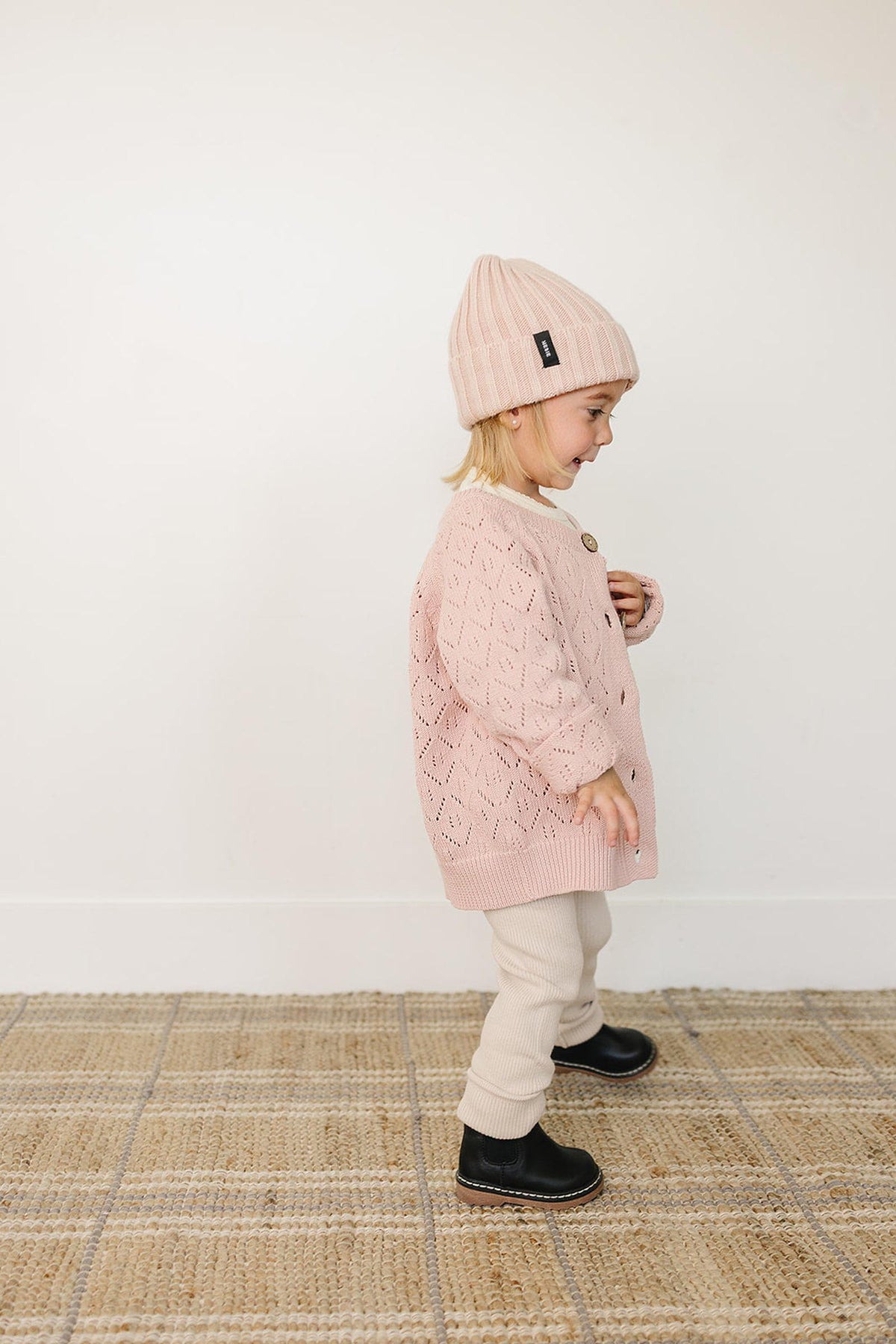 A smiling child wears a light pink chunky knit beanie and a matching sweater, standing on a textured rug.
