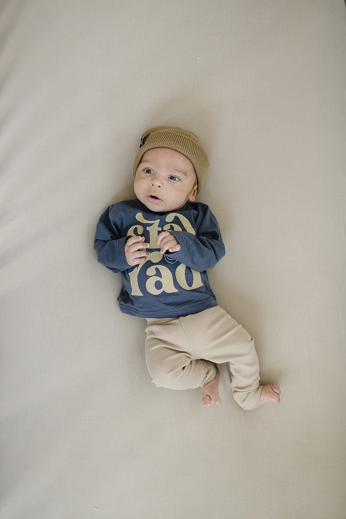 A baby wearing a tan knit hat, blue graphic shirt, and beige ribbed leggings lies on a light fabric.