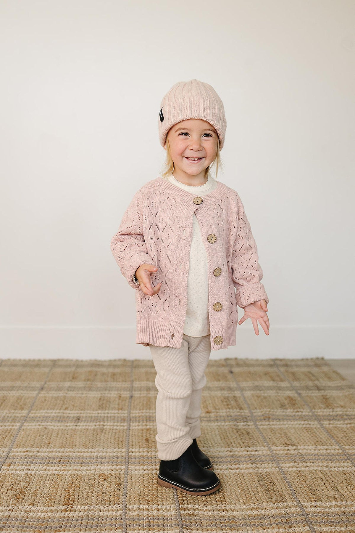 A smiling child wears a light pink chunky knit beanie and matching cardigan, paired with beige pants and black boots.