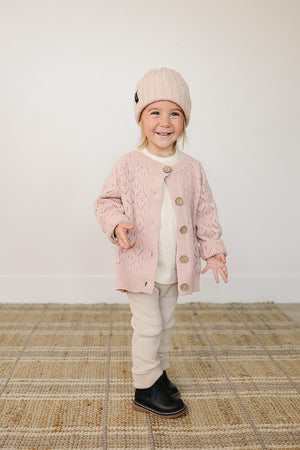 A smiling child wears a light pink chunky knit beanie and matching cardigan, paired with beige pants and black boots.