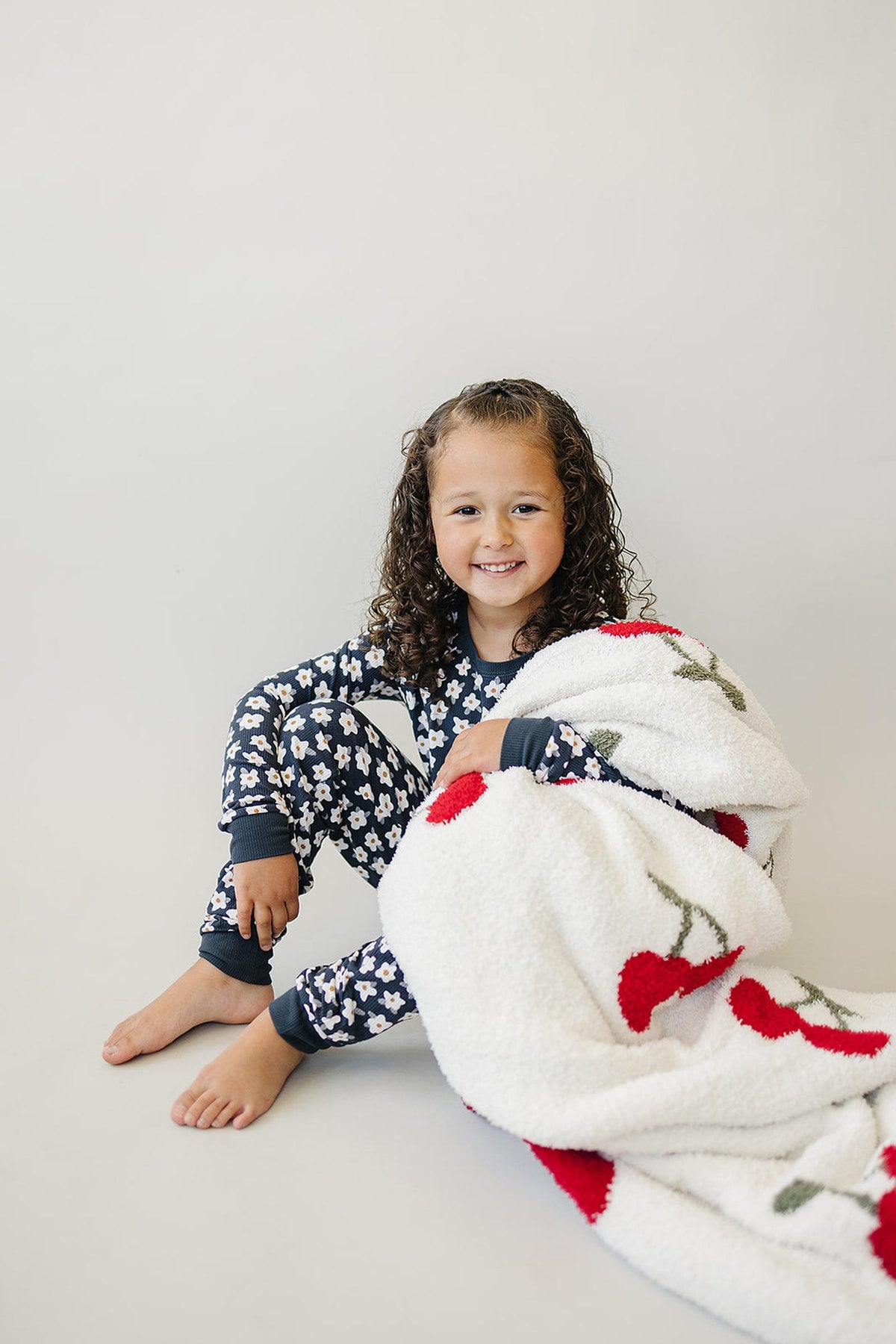 A child in navy daisy-patterned pajamas holds a fluffy white blanket with red cherries, smiling against a plain background.