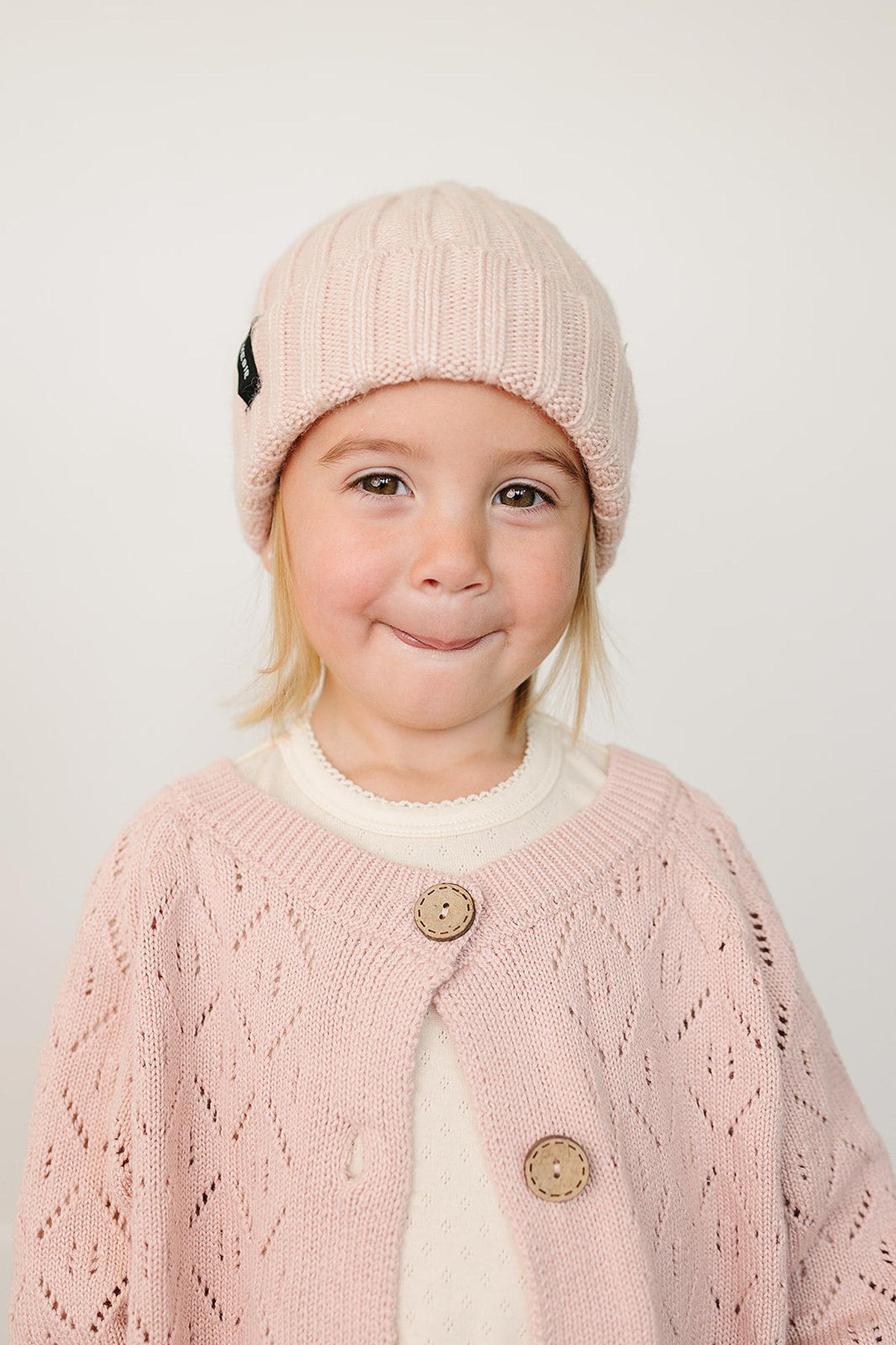 A young child smiles while wearing a light pink chunky knit beanie and a matching pink cardigan.