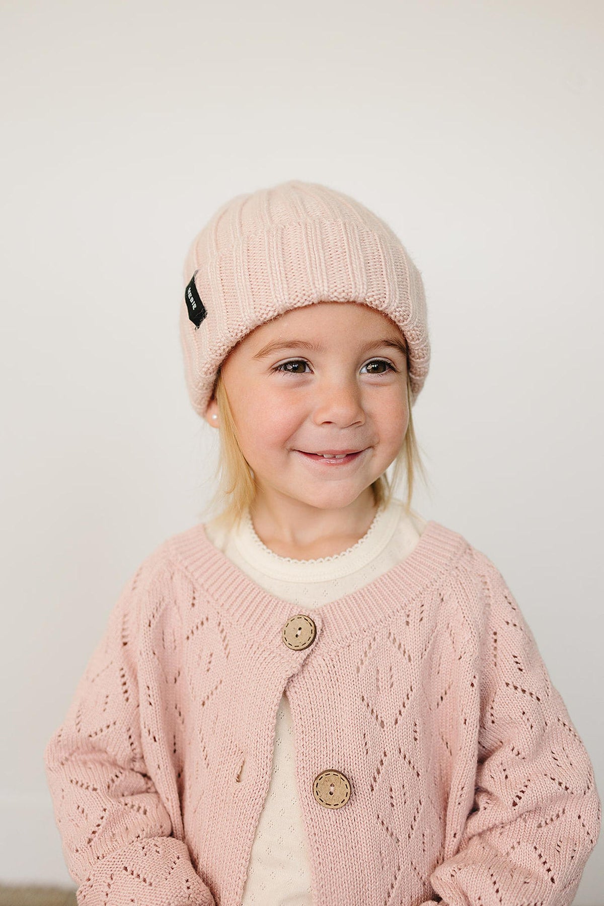 A smiling child wears a light pink chunky knit beanie and a matching cardigan, set against a neutral background.