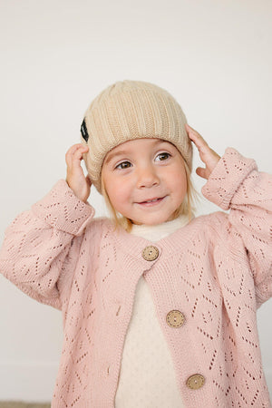 A child wearing a tan chunky knit beanie and a pink patterned cardigan, smiling against a plain background.