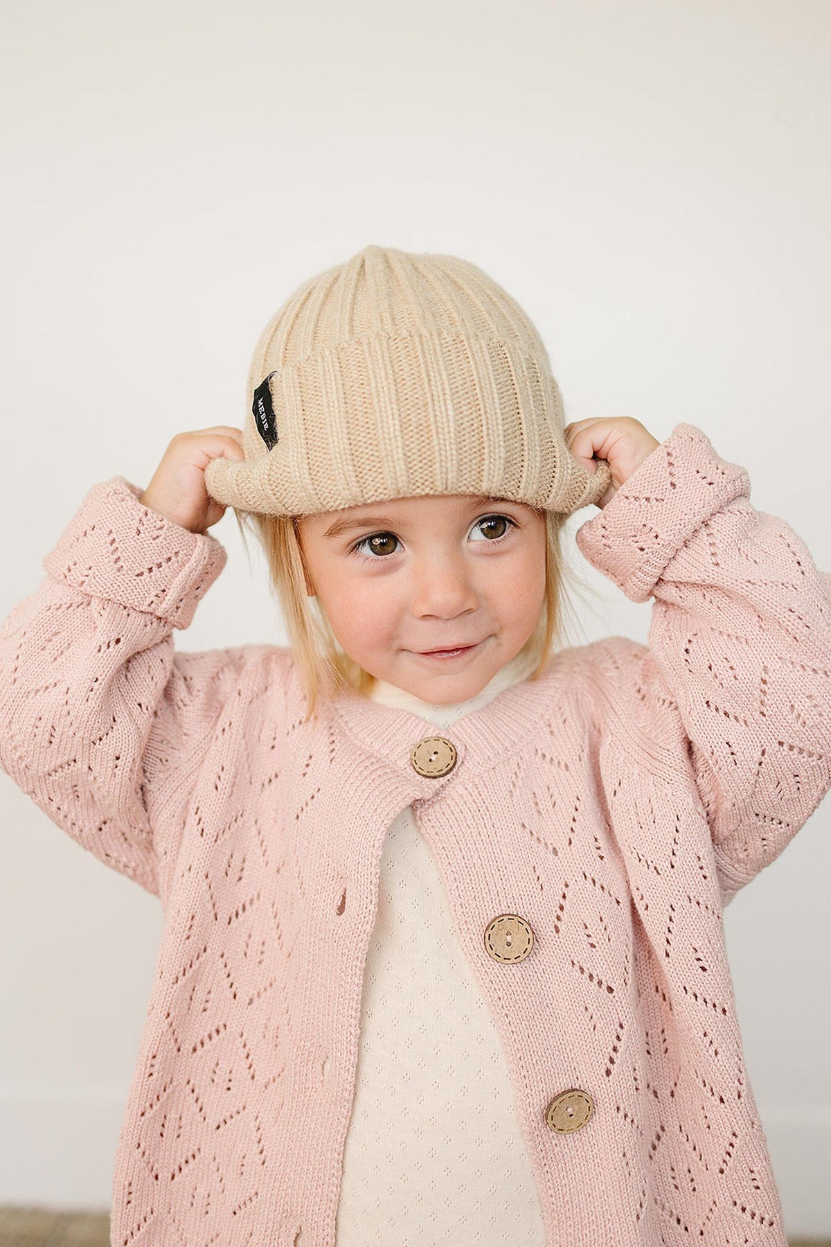 A child wearing a tan chunky knit beanie and a pink knit cardigan with button details.