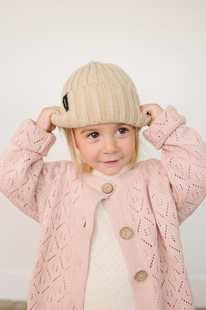 A child wearing a tan chunky knit beanie and a pink knit cardigan with button details.