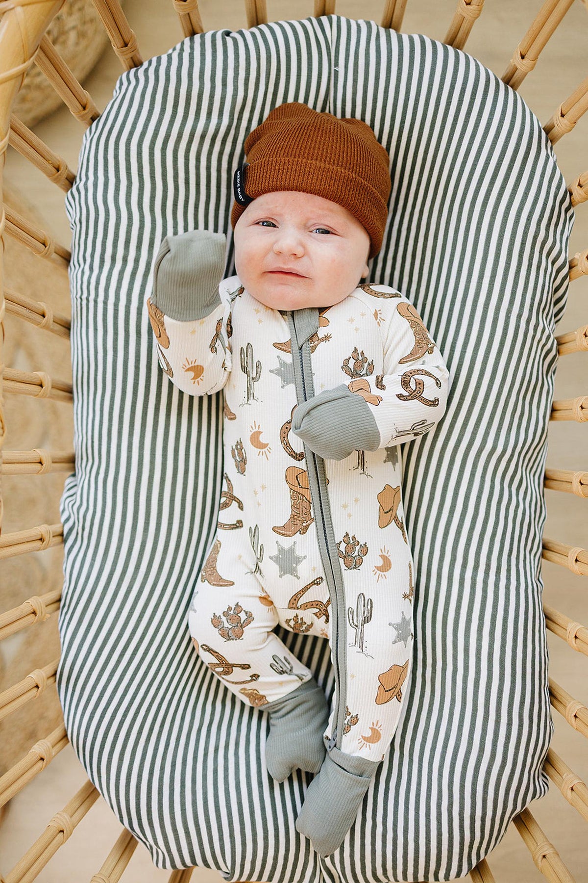 A baby lies in a striped bassinet wearing a ribbed bamboo zipper pajama featuring cowboy and rodeo designs.