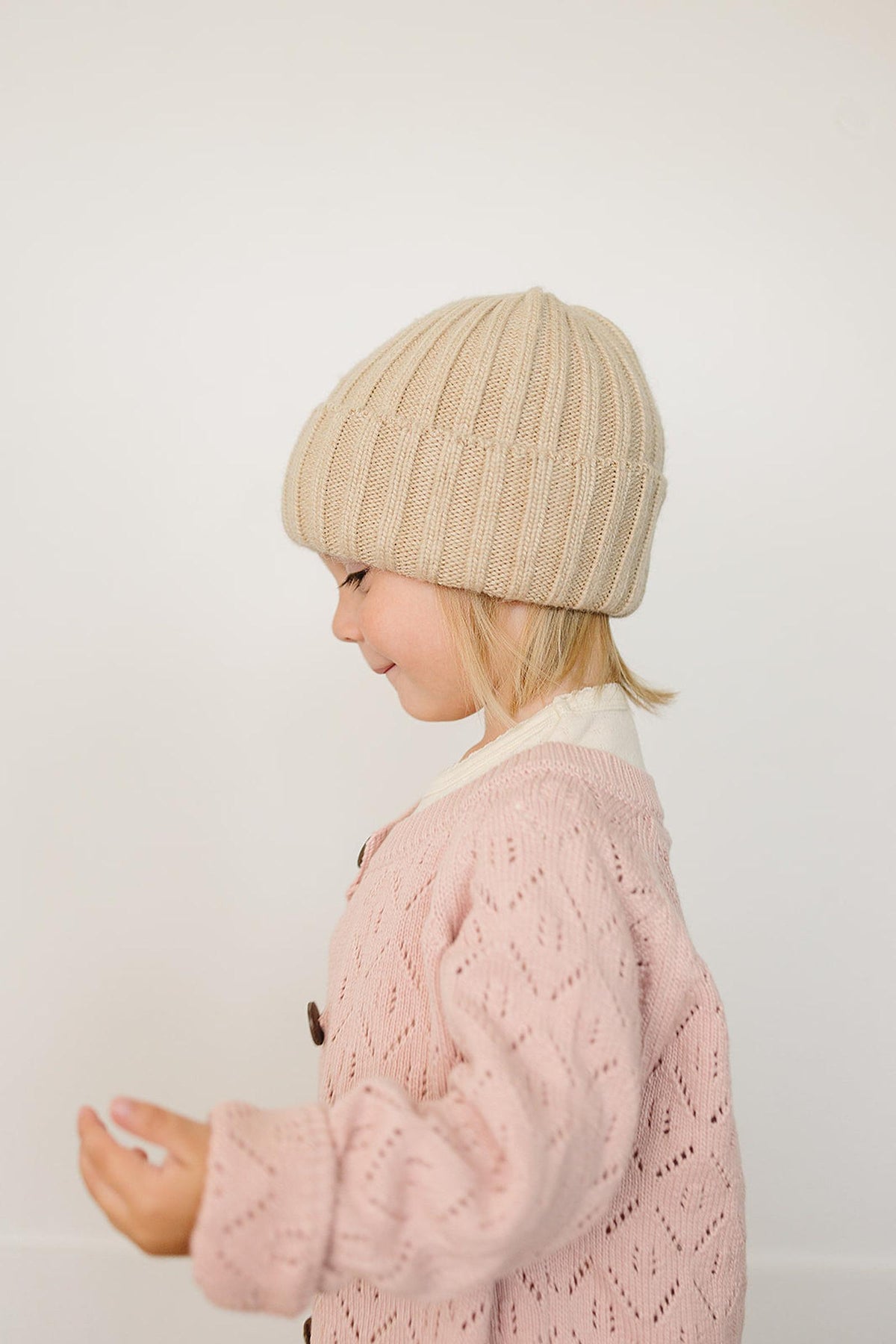 A child in a tan chunky knit beanie and pink sweater stands against a plain white background.