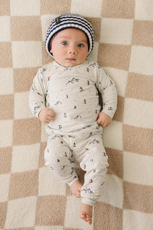 A baby lies on a checkered blanket, wearing a navy and white striped beanie and a patterned pajama set.