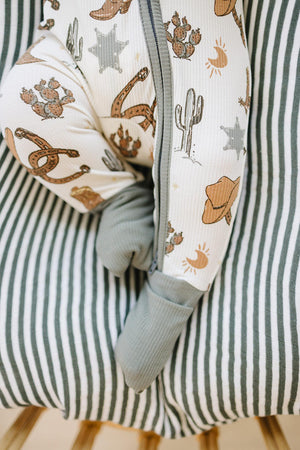 A baby in a cowboy-themed ribbed pajama featuring cacti and horseshoes, with gray footed legs, rests on a striped blanket.