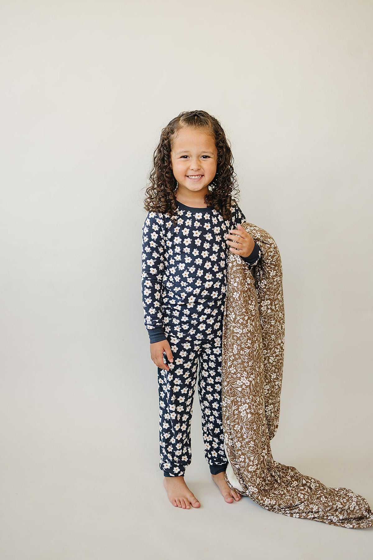 A child wearing a navy pajama set with white daisy patterns holds a patterned brown blanket, smiling.