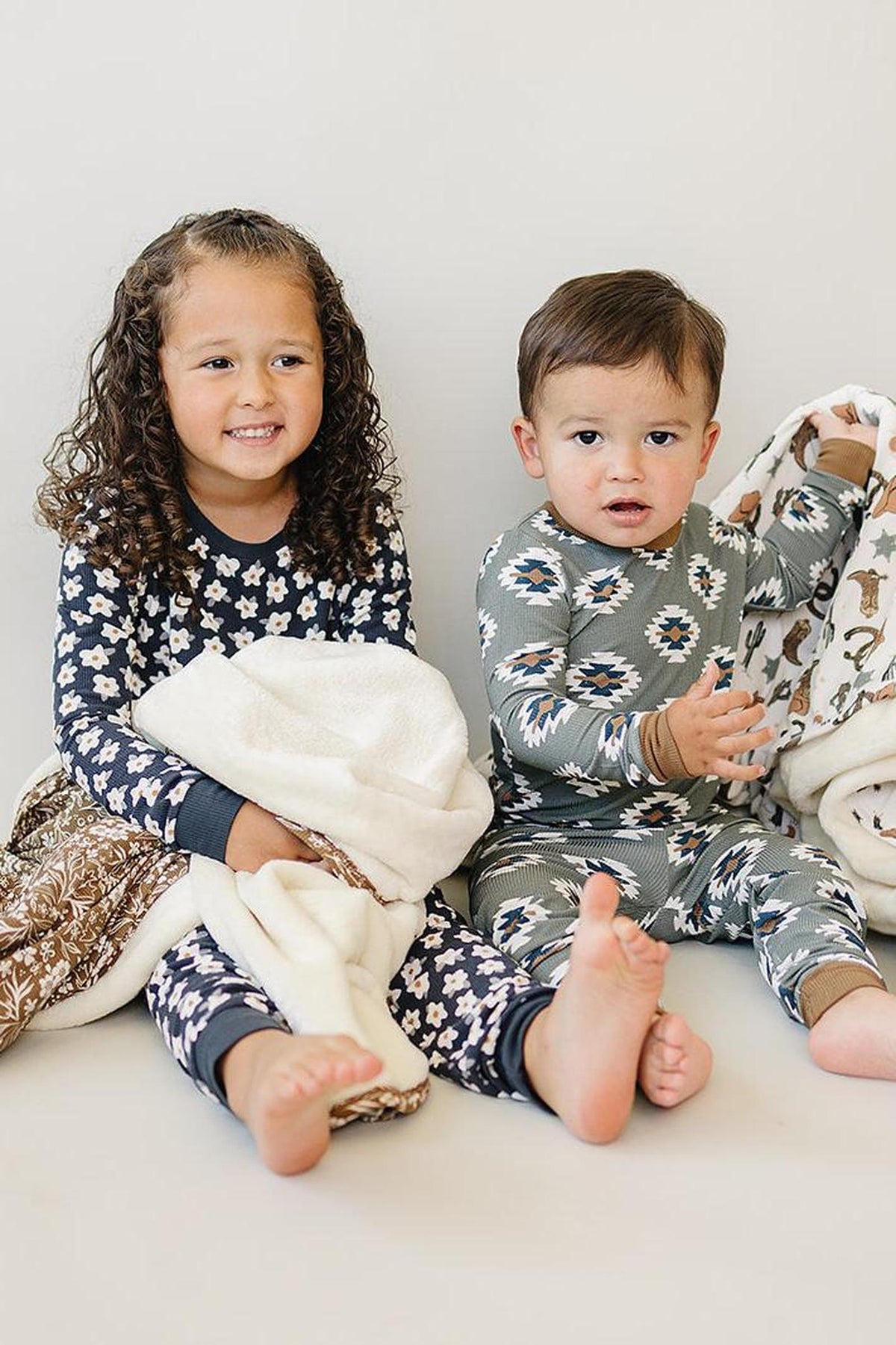 Two children wearing patterned pajamas, one in navy daisy and the other in geometric print, holding blankets.