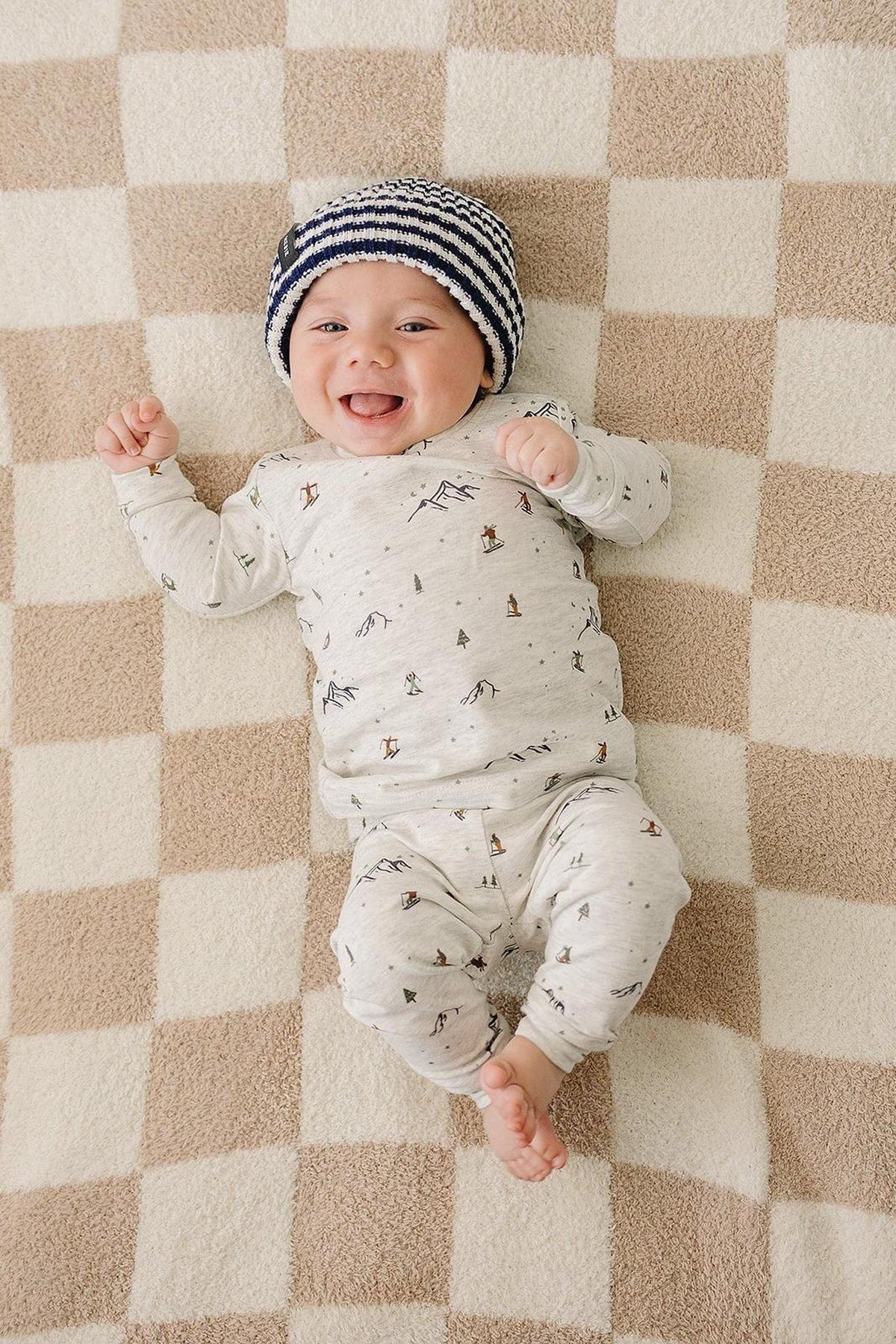A smiling baby lies on a checkered blanket, wearing a navy and white striped knit beanie and a patterned outfit.