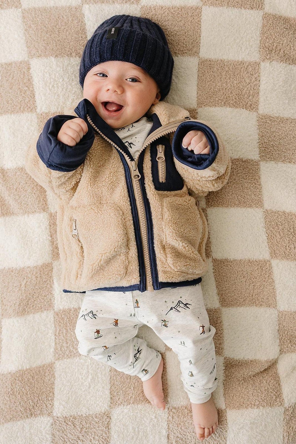 A happy baby wearing a navy knit beanie and a beige fleece jacket lies on a checkered blanket, smiling.