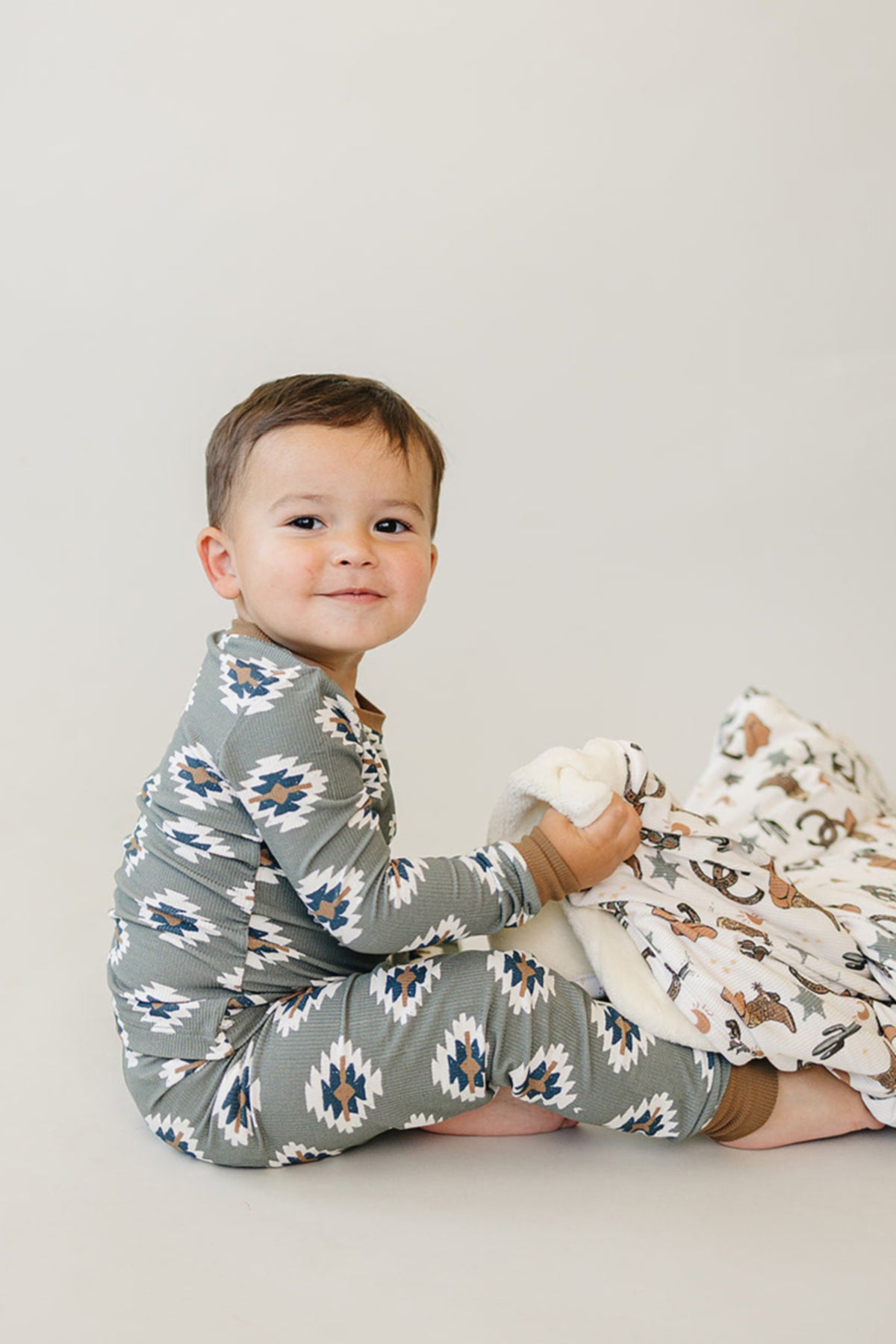 A toddler in a green, Aztec-patterned pajama set sits holding a printed blanket against a plain background.