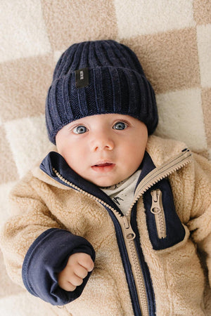 A baby with blue eyes wears a navy chunky knit beanie and a beige fleece jacket, lying on a patterned.