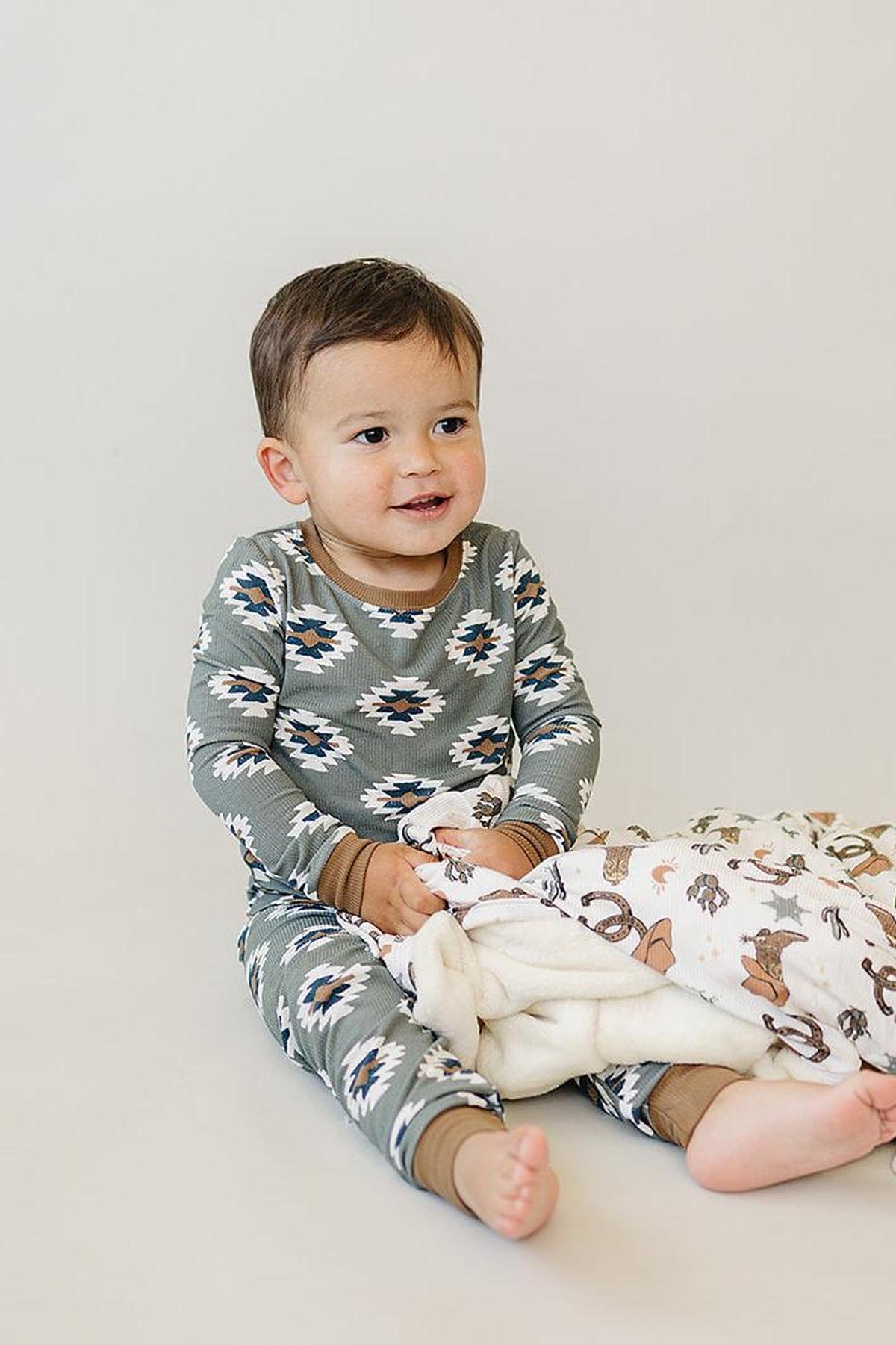 A toddler wearing a green Aztec-patterned pajama set holds a patterned blanket, sitting on a light background.